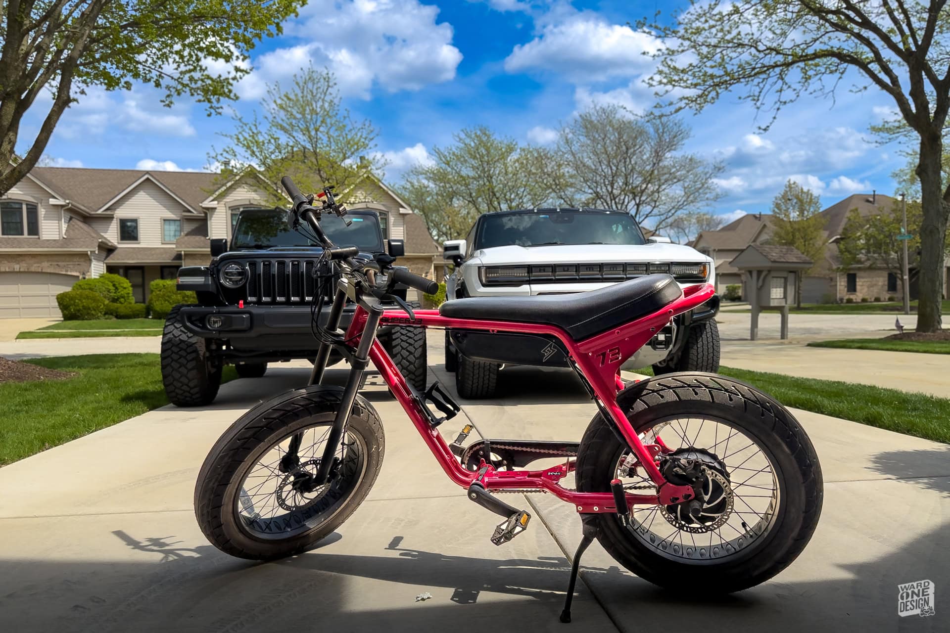 Electric fat tire bike parked on a suburban driveway with two SUVs in the background, surrounded by trees and houses under a bright blue sky.