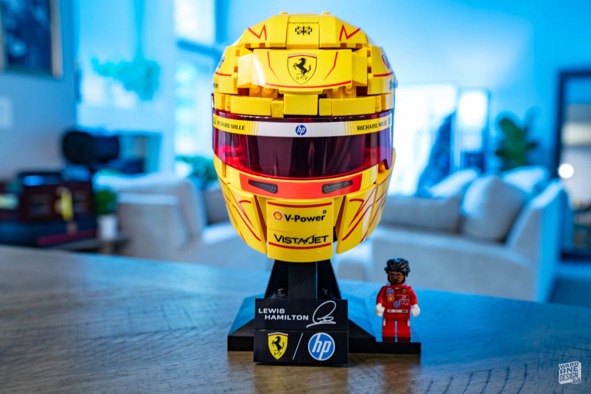 Detailed yellow Ferrari F1 helmet with V-Power and VistAJet logos, displayed on a stand with a small LEGO figure in a red racing suit, in a modern indoor setting.