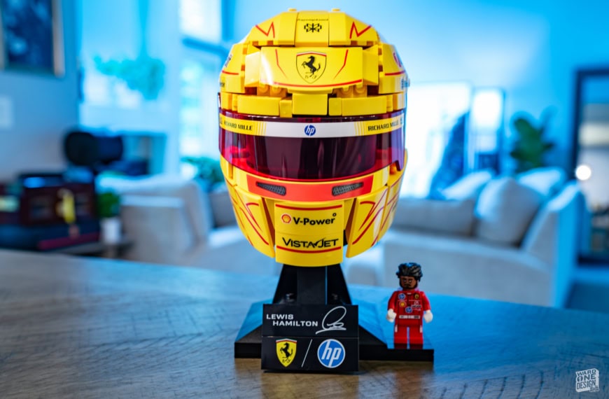 Detailed yellow Ferrari F1 helmet with V-Power and VistAJet logos, displayed on a stand with a small LEGO figure in a red racing suit, in a modern indoor setting.