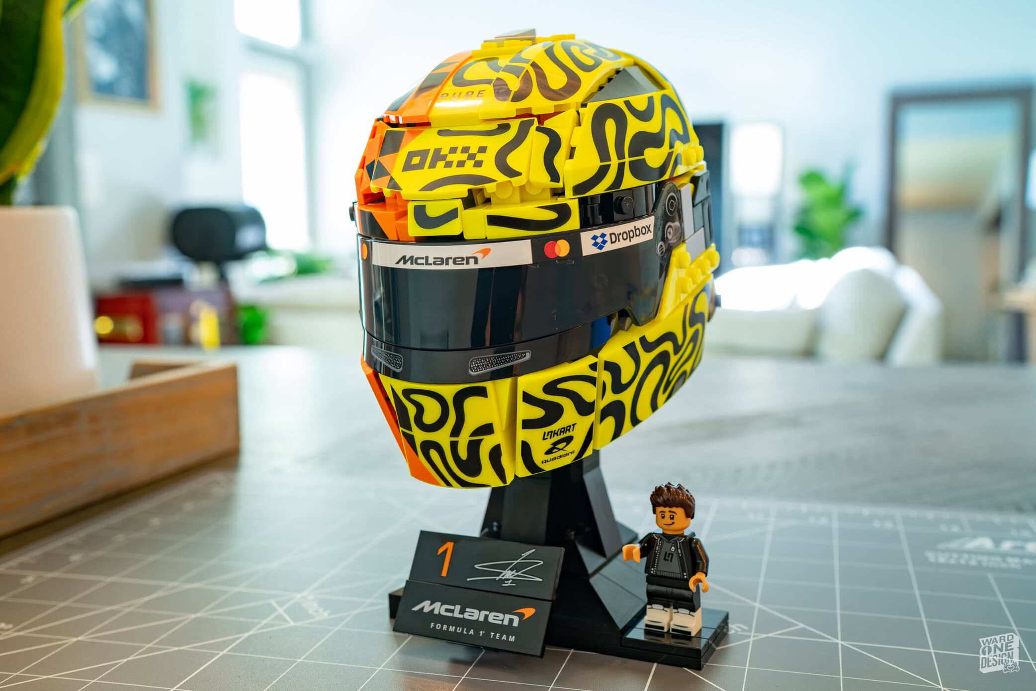 Lego McLaren F1 helmet with McLaren logo and vibrant yellow and black design, displayed on a stand with a small Lego figure in front, in a well-lit room.