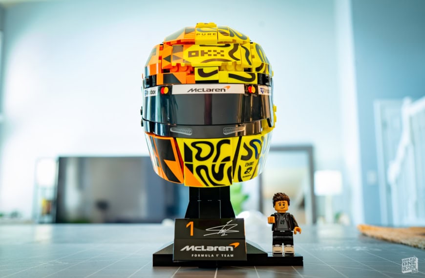 Detailed LEGO model of Lando Norris's McLaren F1 helmet, displayed on a stand with a small LEGO figure in front, in a bright modern room.