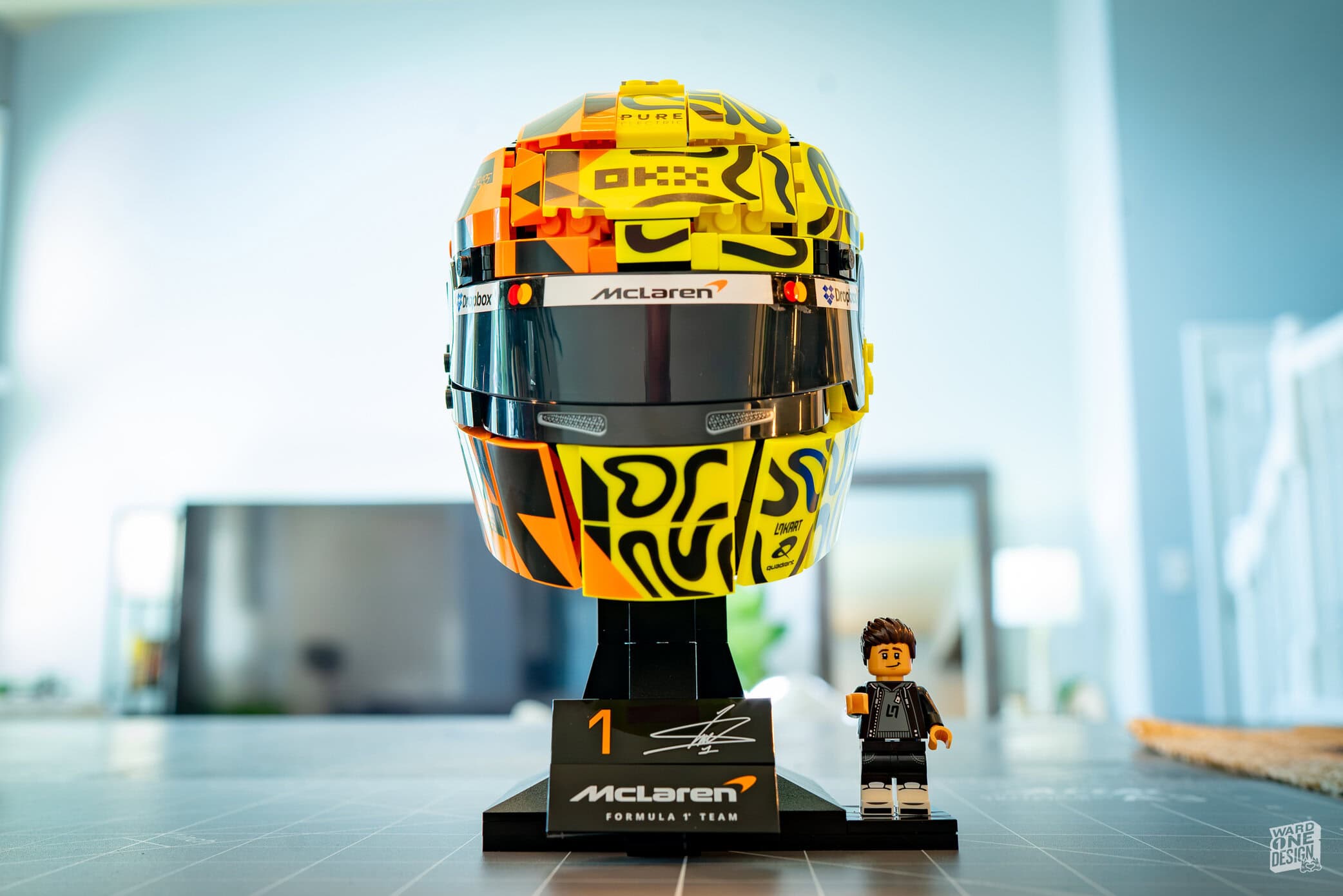 Detailed LEGO model of Lando Norris's McLaren F1 helmet, displayed on a stand with a small LEGO figure in front, in a bright modern room.