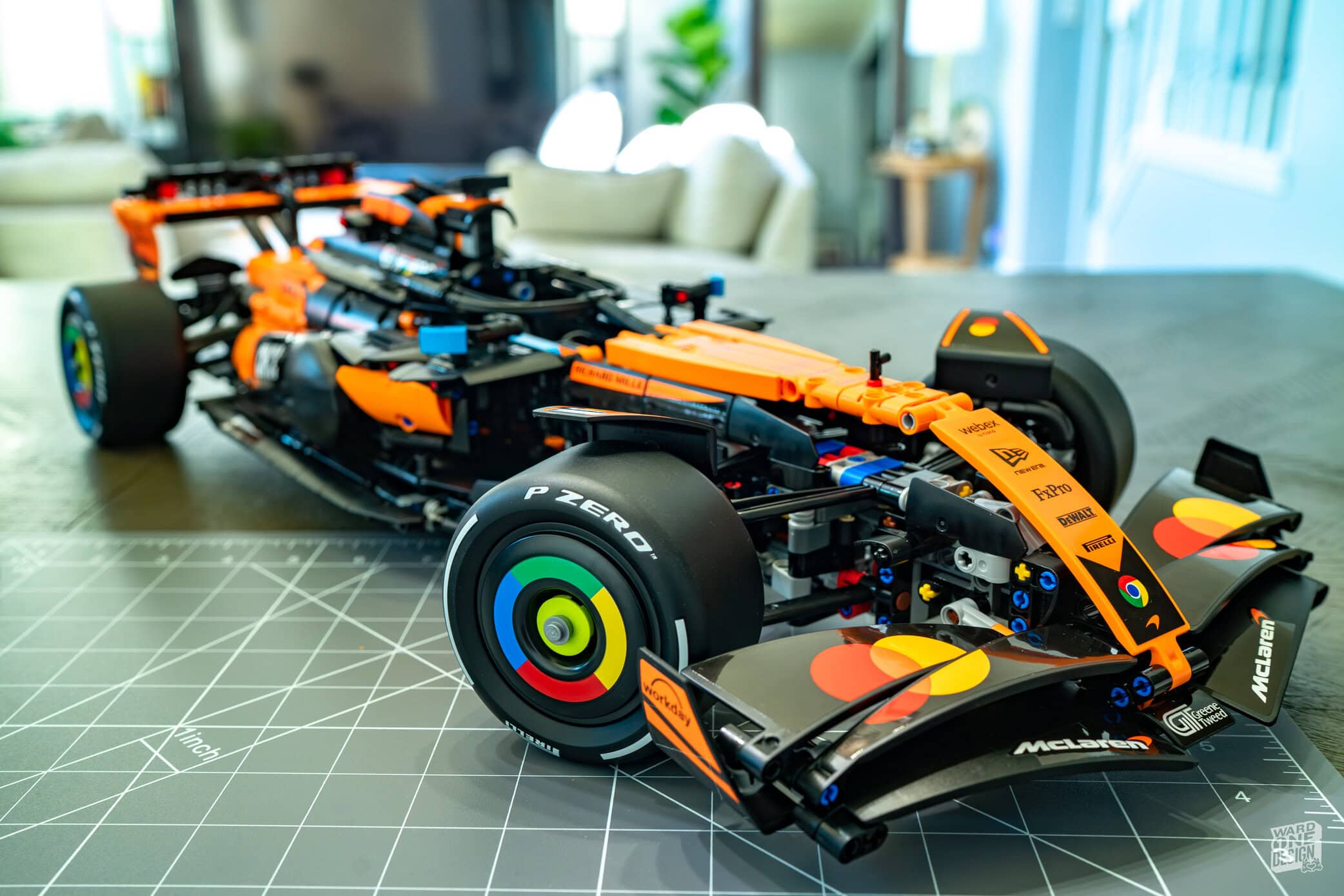 Detailed Lego model of the McLaren MCL39 F1 car in orange and black, displayed on a worktable with a modern living room background.