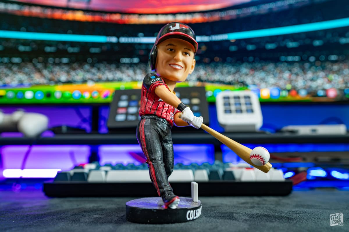 Close-up of a Colson Montgomery White Sox bobblehead figure with a baseball bat, set against a colorful sports event background.