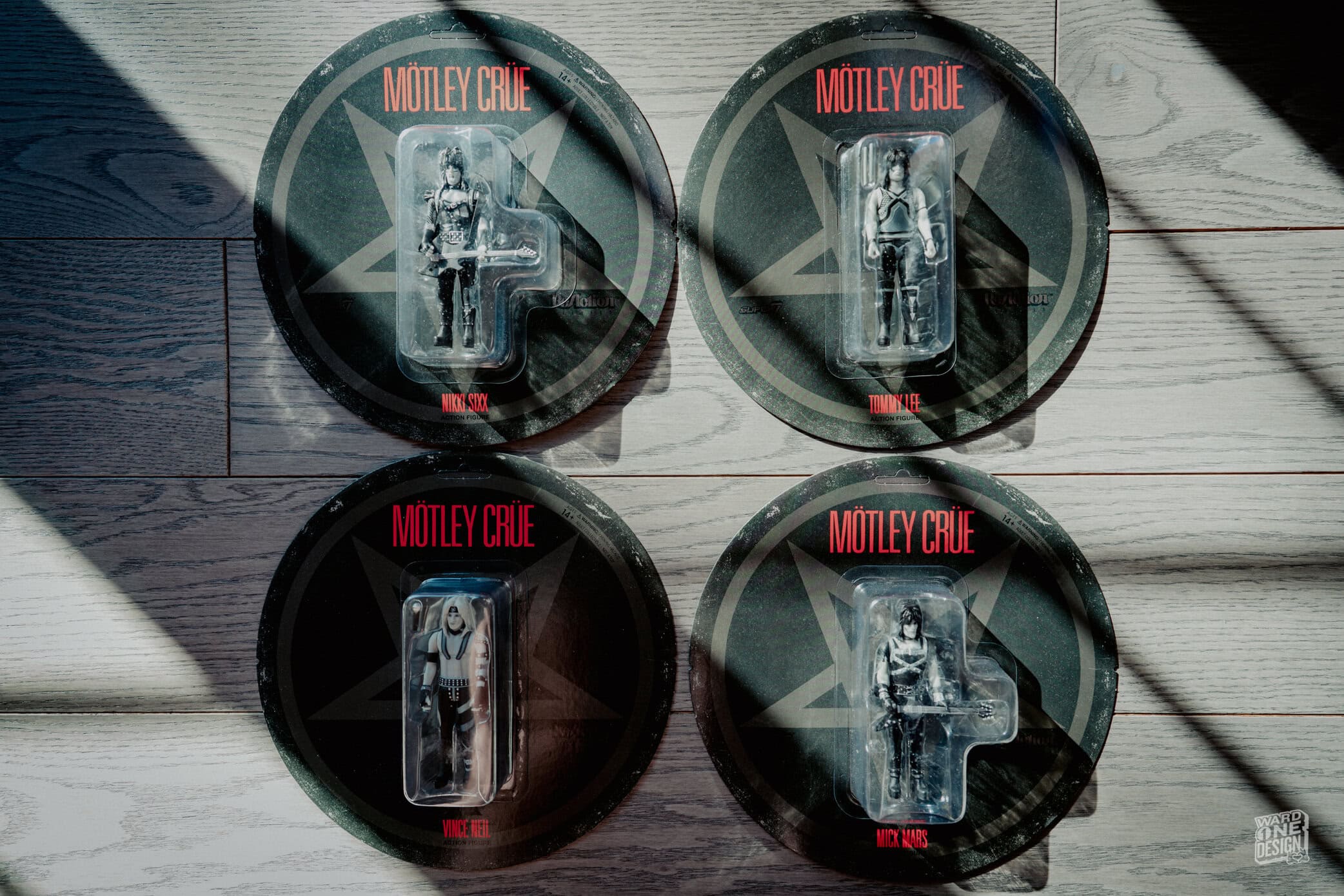 Four Motley Crue Super 7 Wave 2 figures in packaging, featuring band members in detailed designs, displayed on a wooden surface with shadows.