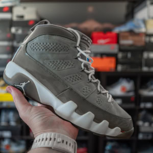 Close-up of Air Jordan 9 Retro Cool Grey sneaker held in hand, showcasing its design and colorway, with a background of sneaker boxes on shelves.