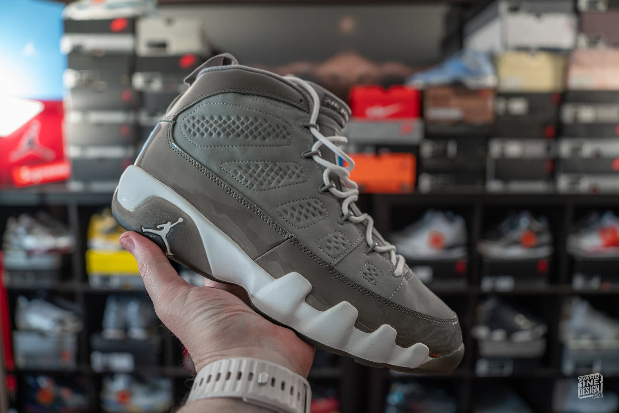 Close-up of Air Jordan 9 Retro Cool Grey sneaker held in hand, showcasing its design and colorway, with a background of sneaker boxes on shelves.