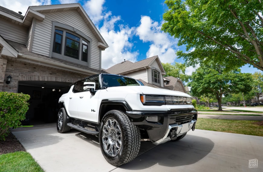 All-electric 2024 Hummer EV SUV parked in a suburban driveway with modern homes and lush greenery in the background.