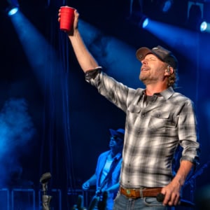 Dierks Bentley on stage during the Broken Branches Tour, holding a red cup and singing passionately with a band in the background, under vibrant concert lighting.