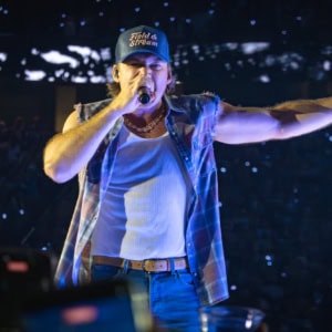 Morgan Wallen singing passionately with a microphone, wearing a baseball cap and casual attire, with concert lighting and audience in the background.