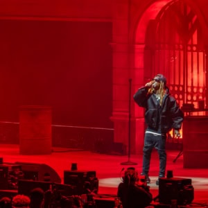 Lil Wayne on stage during Tha Carter VI Tour, illuminated by red lighting, performing passionately with a microphone in hand at a concert venue.